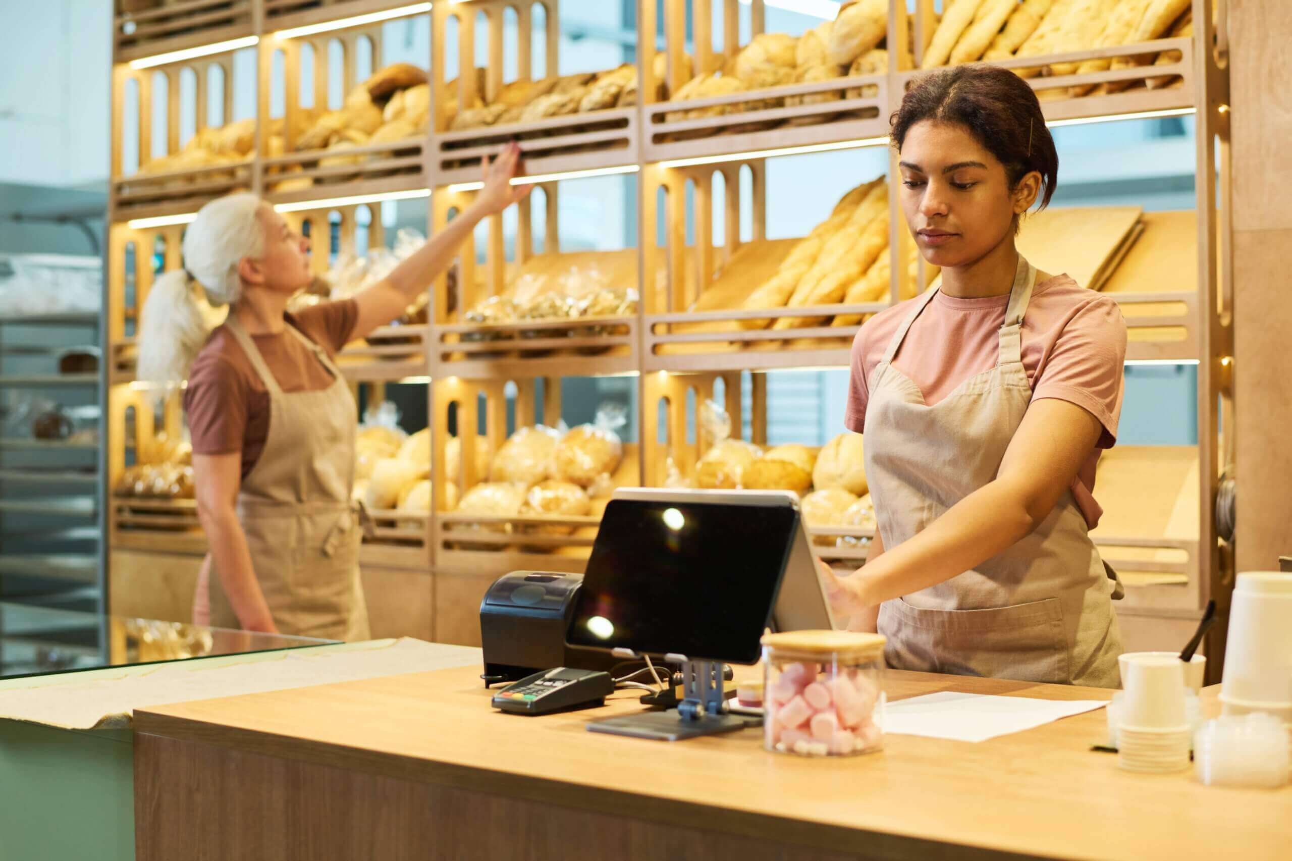 Two bakery employees at work: one standing at the counter operating a POS system, and the other arranging bread on wooden shelves filled with various types of baked goods in the background