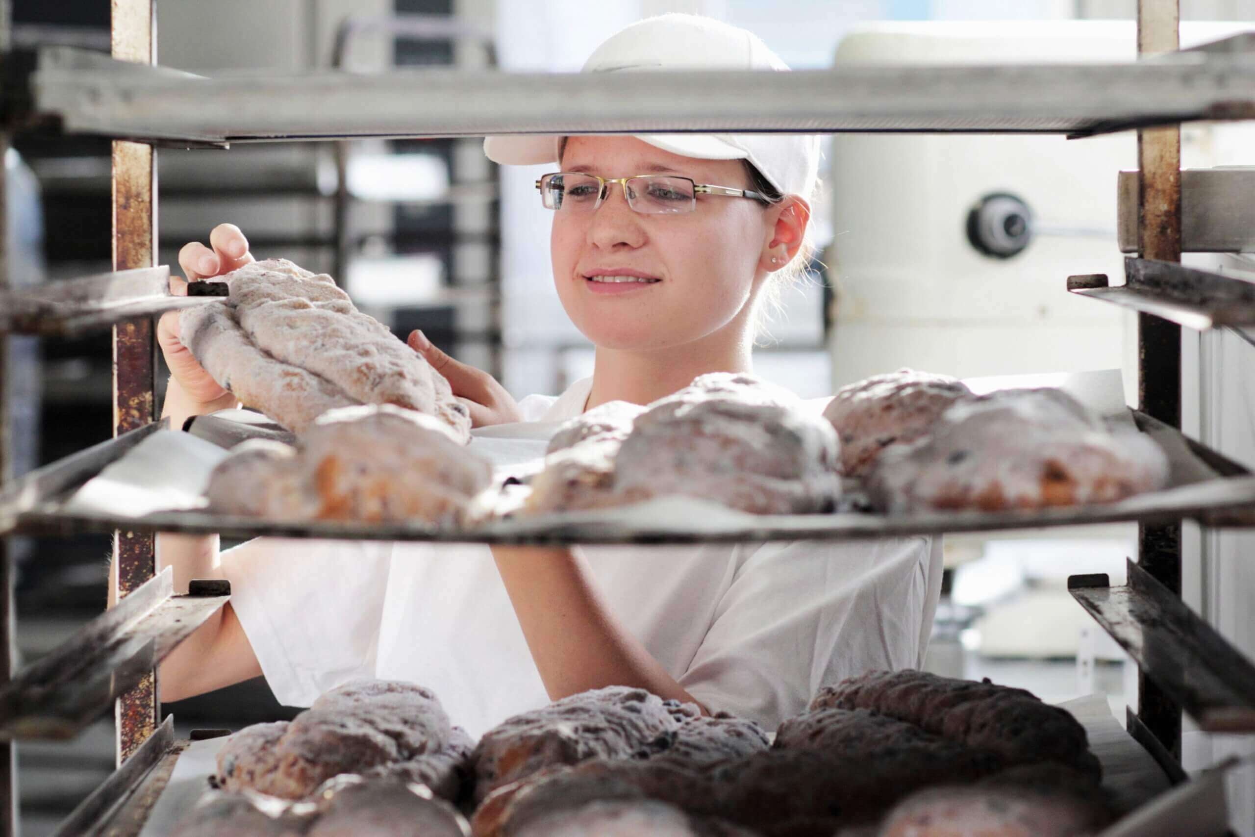 A bakery worker wearing a white uniform and cap carefully inspecting a loaf of powdered bread on a metal rack filled with freshly baked goods