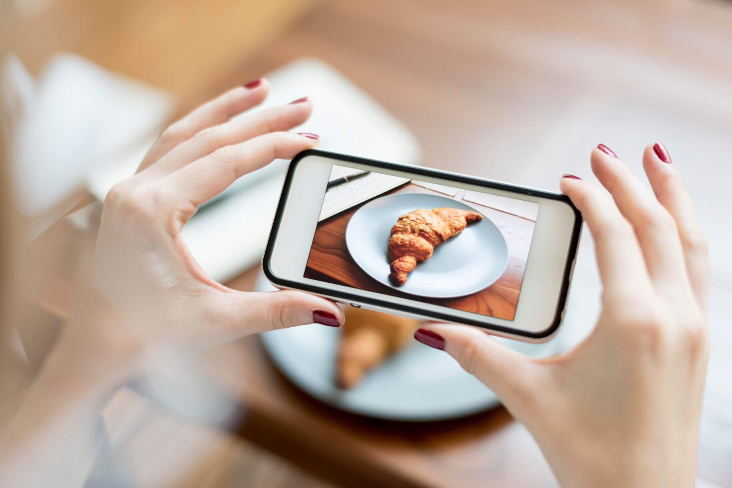 A woman taking a photo of a croissant on a plate with her smartphone