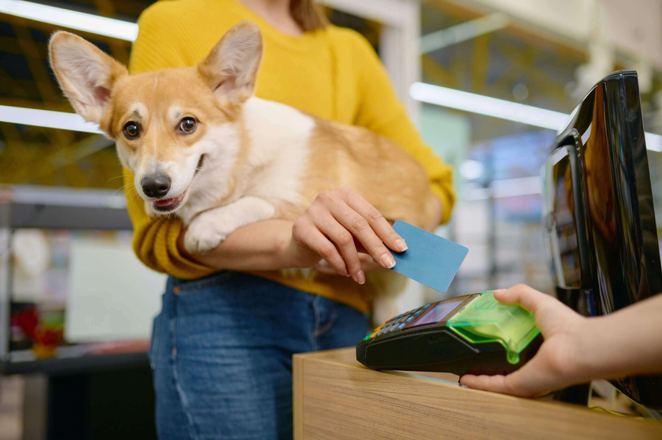 A woman paying for her dog at a pet store checkout counter. She is holding her dog in her arms and swiping a credit card through a card reader