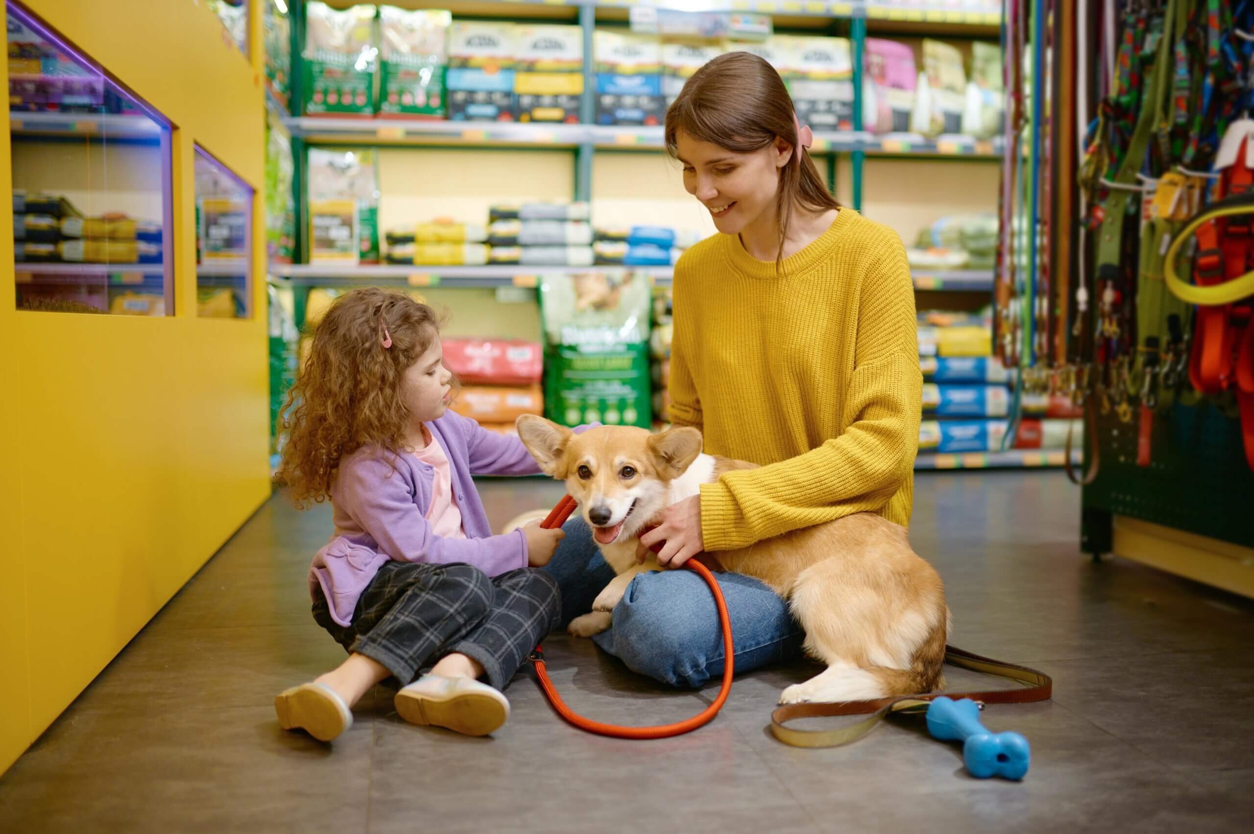 A woman and a young girl are seated on the floor, playing with a dog, highlighting pet store loyalty programs benefits