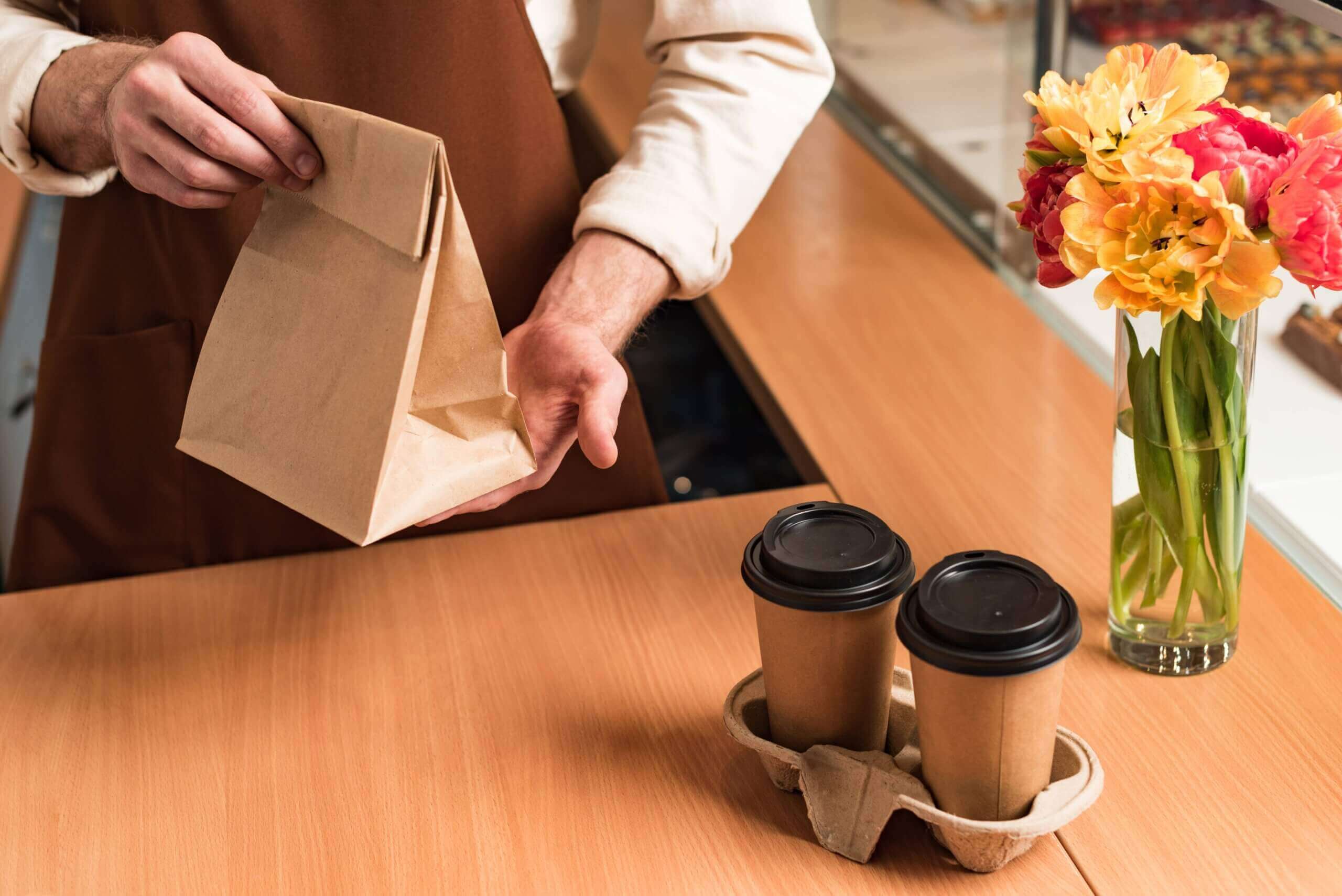 Partial view of barista in brown apron with paper bag and paper cup of coffee to go