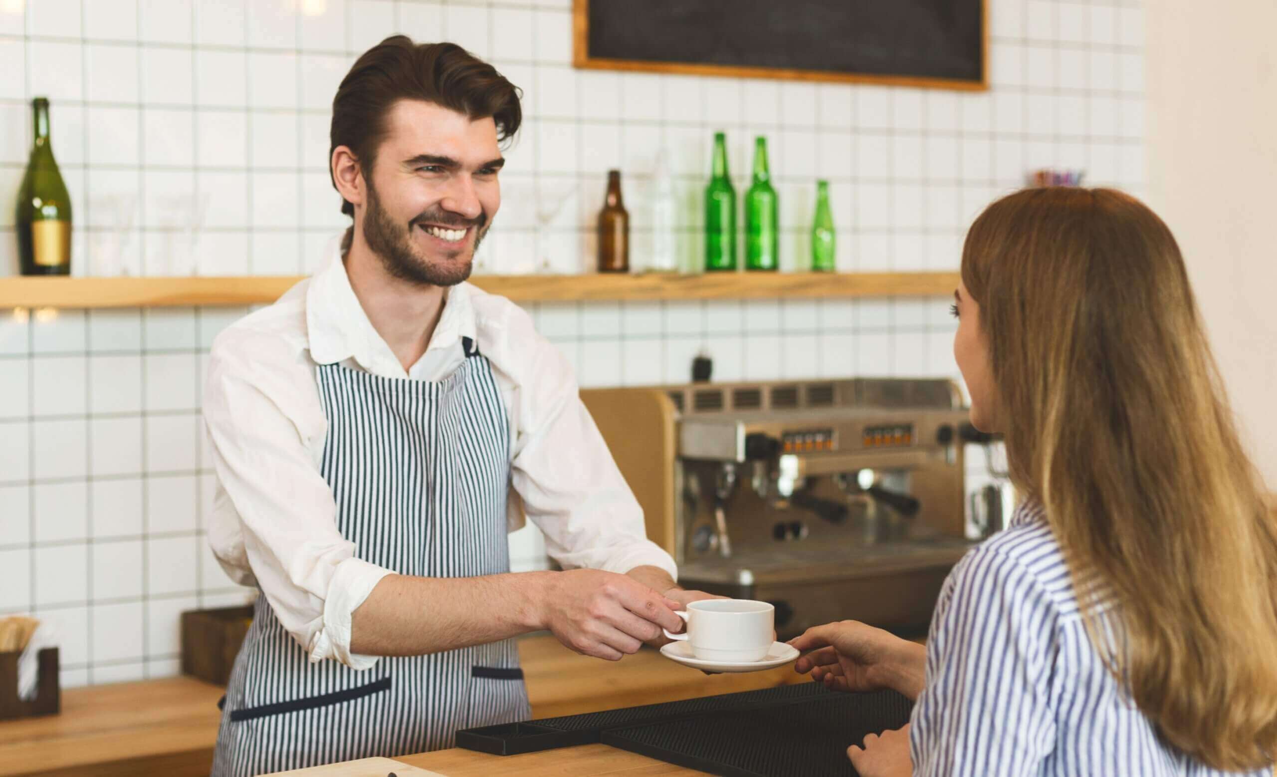 A happy barista smiling and offering a coffee mug to a customer at the counter
